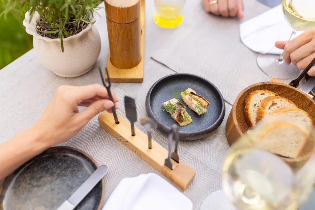 Ein eleganter Tisch mit einem kleinen Teller fein angerichteter Speisen, altem Holz und frischem Brot. Grüne Pflanze im Hintergrund.