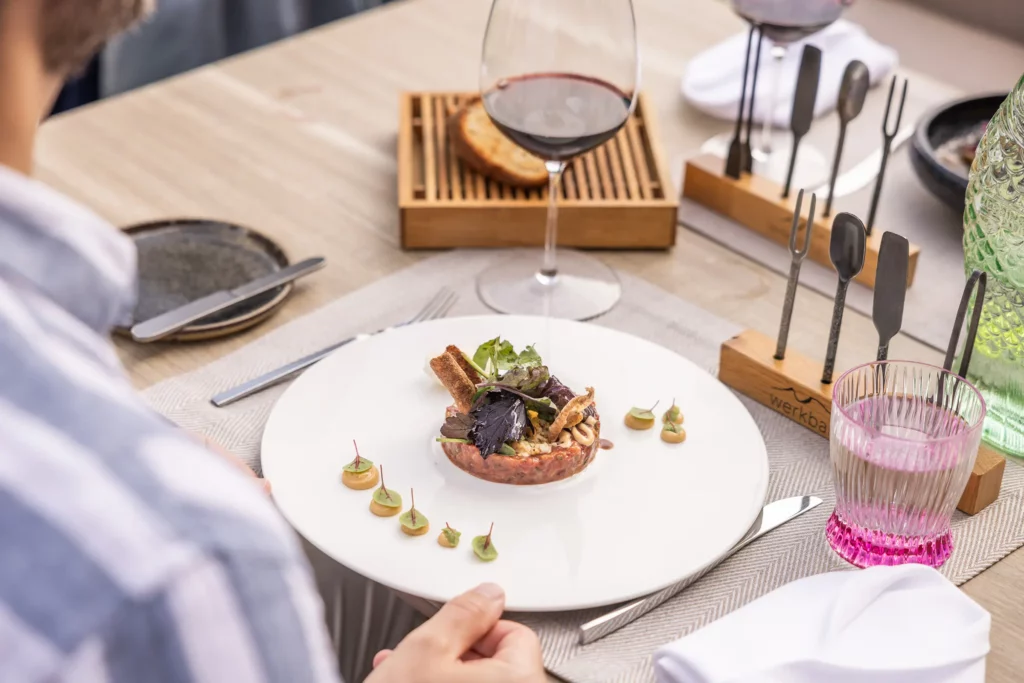 Ein elegantes Gericht auf einem weißen Teller, garniert mit frischen Kräutern und kreativen Beilagen. Rotwein und rosa Wasser in stilvollen Gläsern.