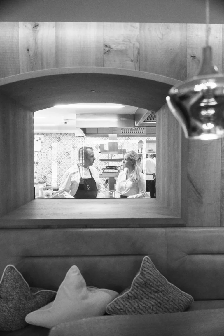 Zwei Personen im Gespräch, durch einen weichen, holzrahmigen Durchgang sichtbar. Im Hintergrund eine moderne Küche mit floralen Fliesen.