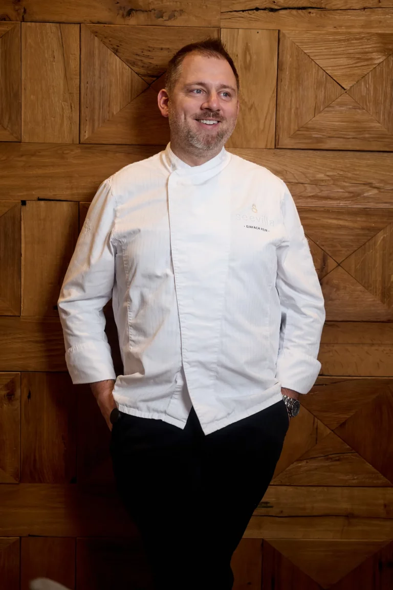 Lächelnder Koch in weißem Chefkoch-Outfit vor einer warmen, rustikalen Holzvertäfelung, entspannt mit einer Hand in der Hosentasche.