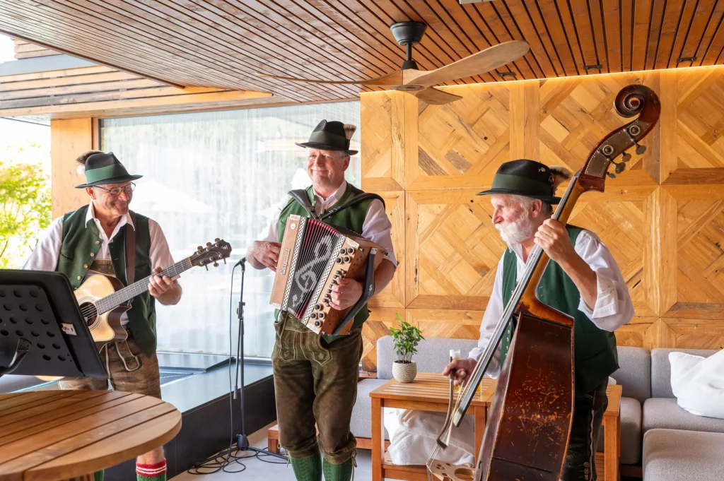 Drei Musiker in traditioneller Tracht spielen Akkordeon, Gitarre und Kontrabass in einem stilvollen, hellen Holzraum.