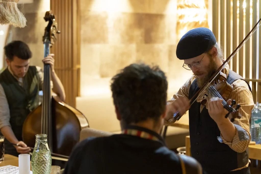 Zwei Musiker spielen in einem eleganten Restaurant: Ein Geiger mit Bart und Brille sowie ein Kontrabassist im Hintergrund. Warme Farben dominieren.