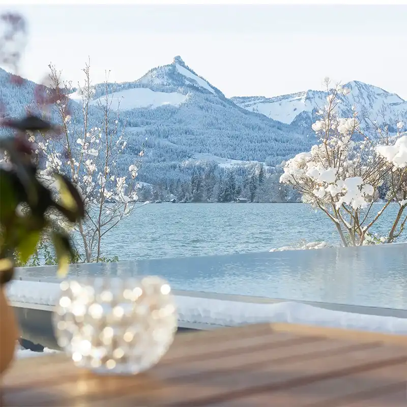 Blick auf den schneebedeckten Berg und den Wolfgangsee, umrahmt von winterlichen Pflanzen und einer glitzernden Glasschale.